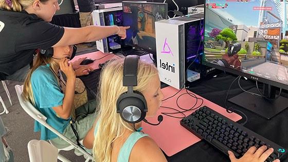 两个小女孩坐在台式电脑前玩《我的世界》(Minecraft)，一个穿着Super Girl Gamer Pro衬衫的女人在旁边帮助她们.