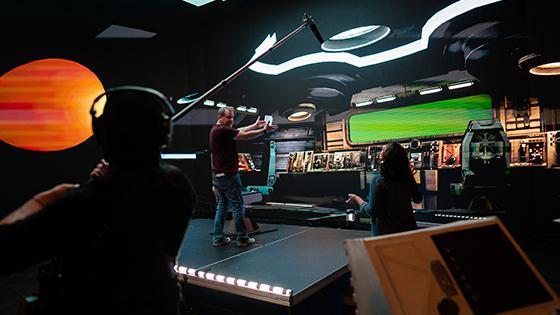 Space Pups’ director Jason Murphy stands onstage in a virtual spaceship set. He is talking to a crew member while another crew member holds a boom mic.