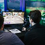 Two people have their backs to the camera while watching ‘NBA 2K’ gameplay on a small monitor on a table in front of them.