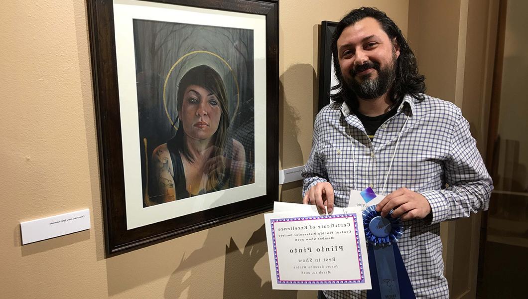 一个微笑的男人站在一个女人的水彩画肖像旁边. 他拿着蓝丝带和最佳表演奖证书.