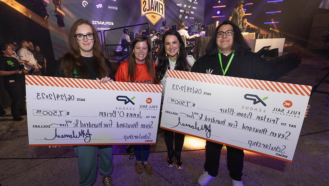Four people standing to the side of a stage while holding large novelty checks and smiling.
